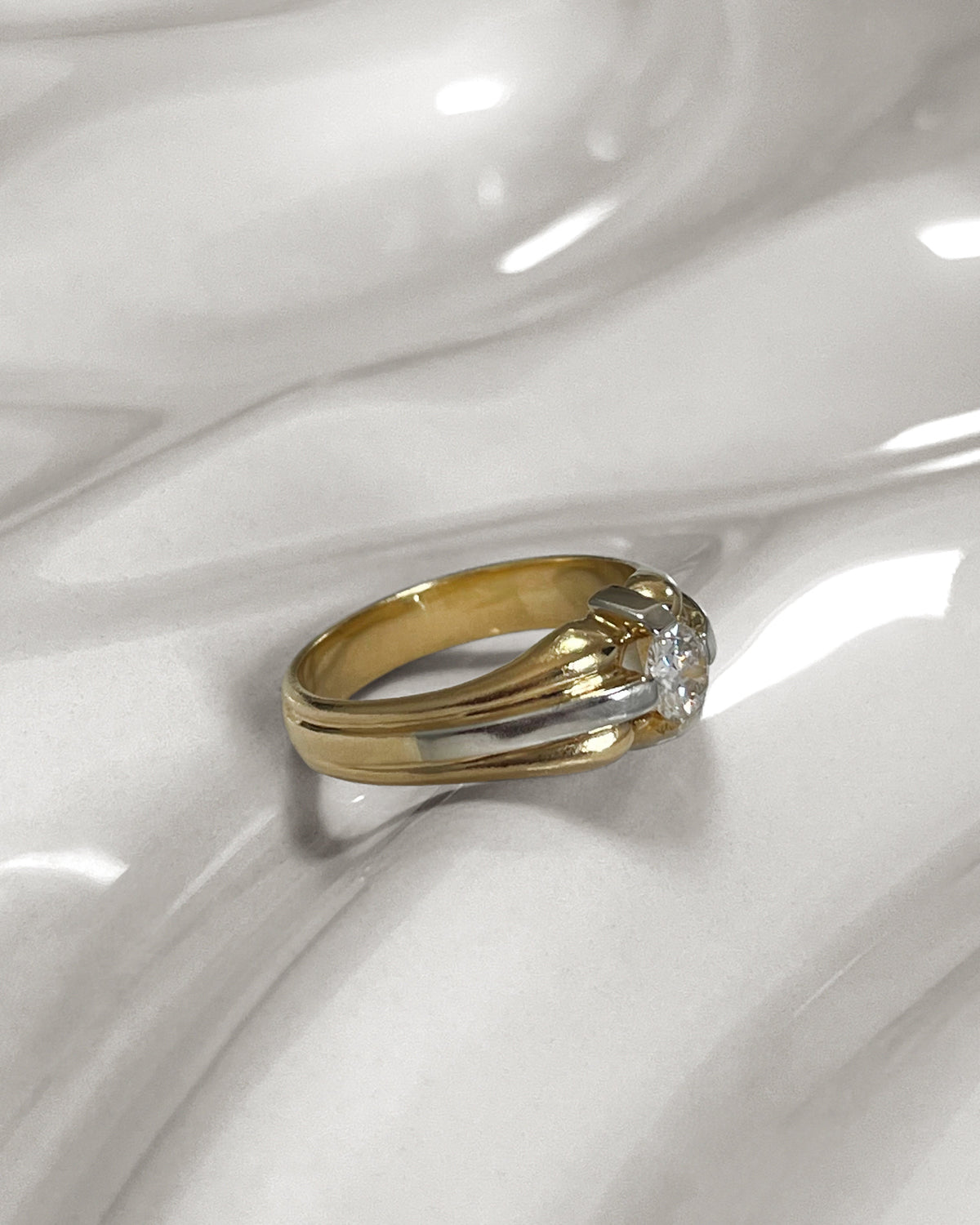 Side view of a 1990s vintage 18kt two-tone gold diamond solitaire engagement ring with a 0.20ct Old European cut diamond in a compass setting, shown on an undulating white tile.