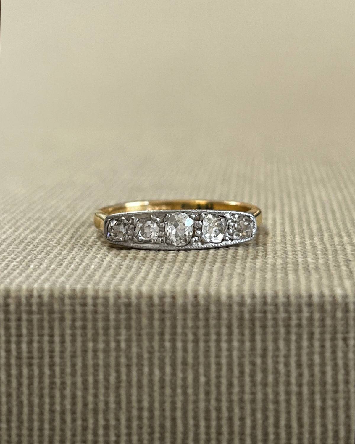 Antique five-stone diamond ring featuring five graduating cushion-shaped Old Mine cut diamonds in delicate millegrain platinum settings, shown against a beige canvas background.