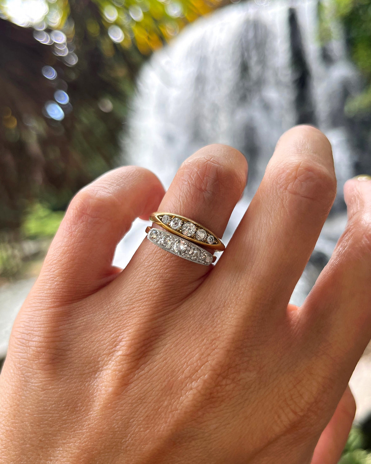 Antique Victorian 18kt gold navette diamond ring shown on a hand beside a five-stone diamond ring, with a waterfall in the background.