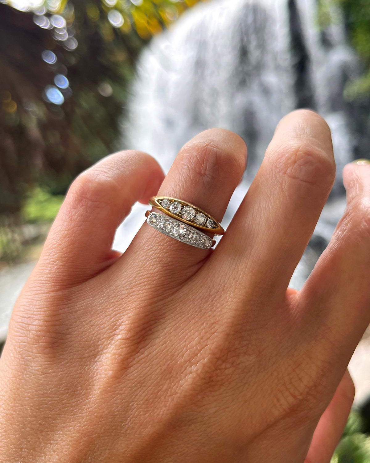 Antique five-stone Old Mine cut diamond ring stacked beside a diamond navette ring, shown on a hand with a waterfall in the background.