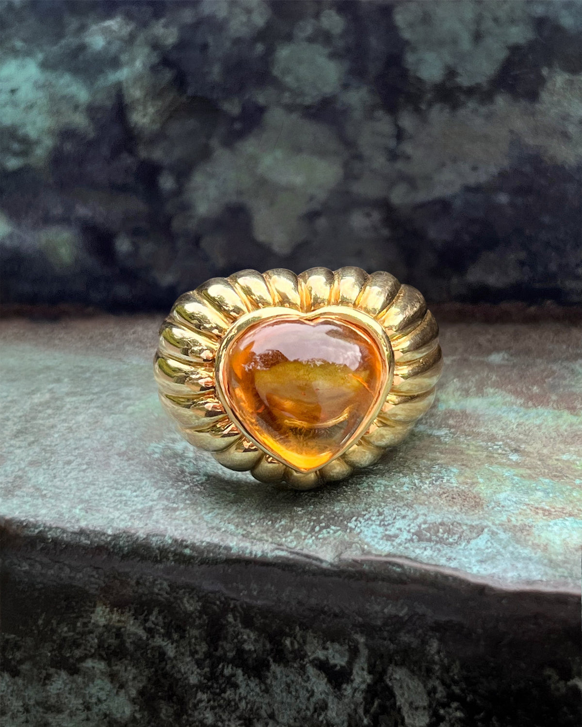 Vintage chunky 18kt gold statement ring with a heart-shaped golden citrine cabochon and ribbed gold detailing, shown against a green bronze background.
