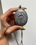 Antique French Art Nouveau silver iris pocket mirror locket held in a hand, suspended from a substantial oval bale with a silver chain, against a white wall.