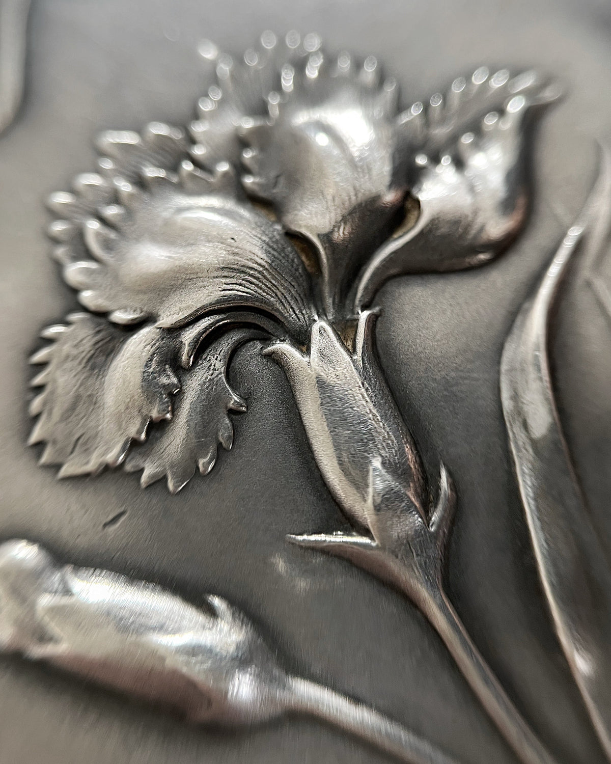 Magnified close-up of the chased iris flower decoration on an antique French Art Nouveau silver pocket mirror locket, showing the elongated petals and naturalistic scrolling border.
