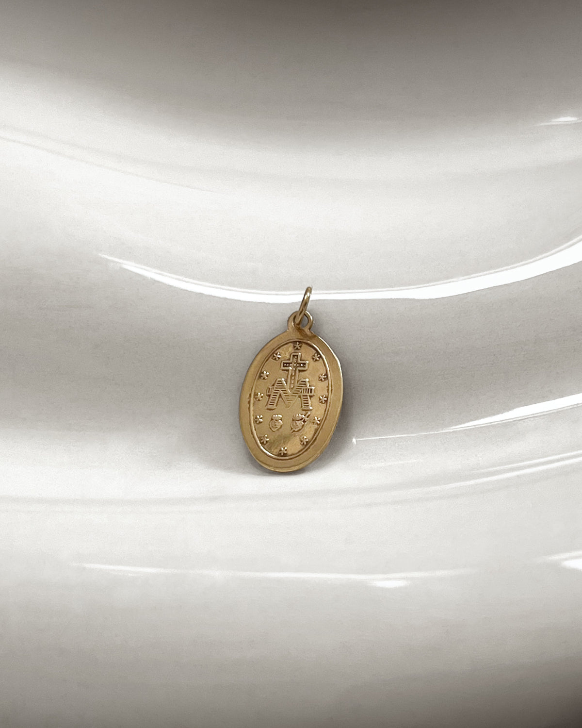 Reverse of a vintage 9kt yellow gold Miraculous Medal pendant on an undulating white tile, showing the traditional M and cross with Sacred Heart and Immaculate Heart symbols.