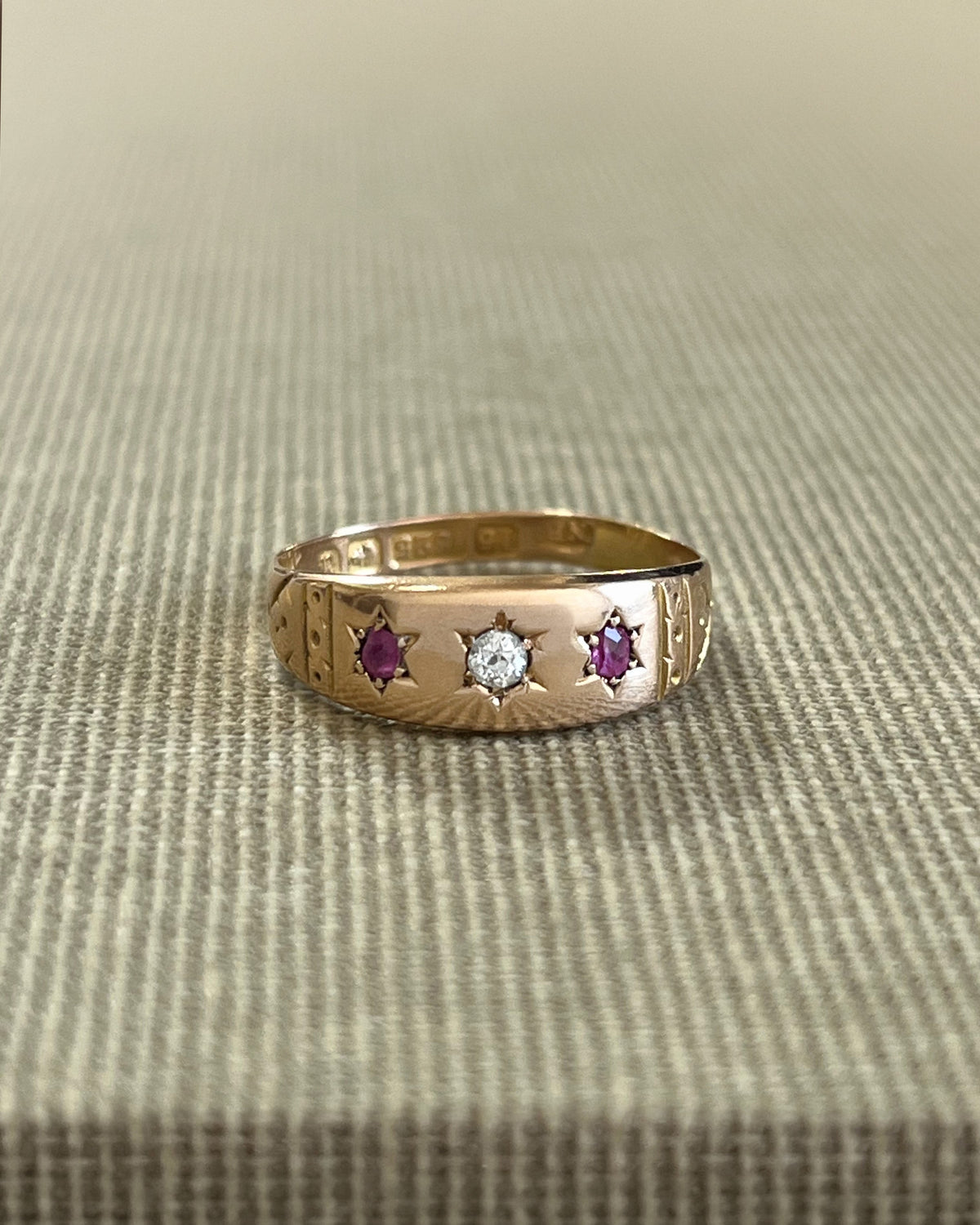 Antique 15kt gold gypsy set ring featuring a diamond and two rubies in six-rayed star settings, with engraved shoulders, shown against a beige canvas background.