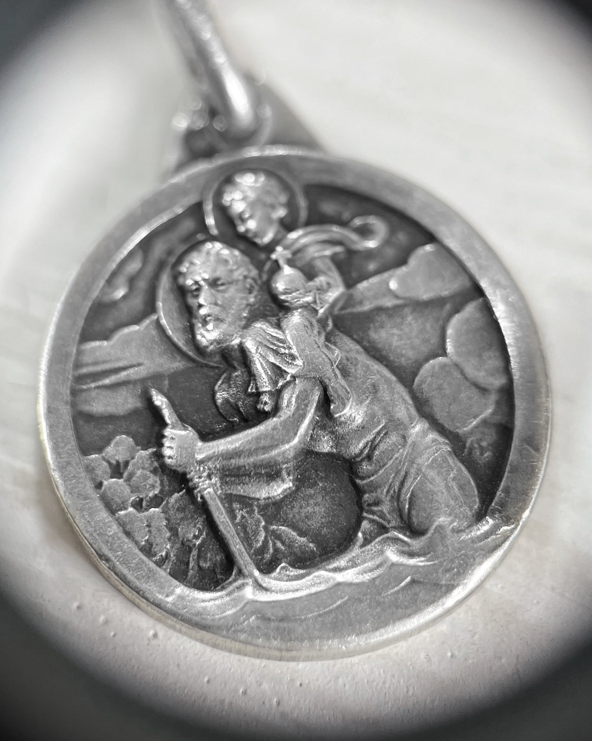 Close-up of 1960s French vintage silver St Christopher pendant showing detailed figural relief and raised border beneath a jeweller's loupe