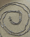 Belle Époque French antique silver sautoir necklace with decorative tubular, marquise and belcher links, arranged in a spiral on textured beige canvas.