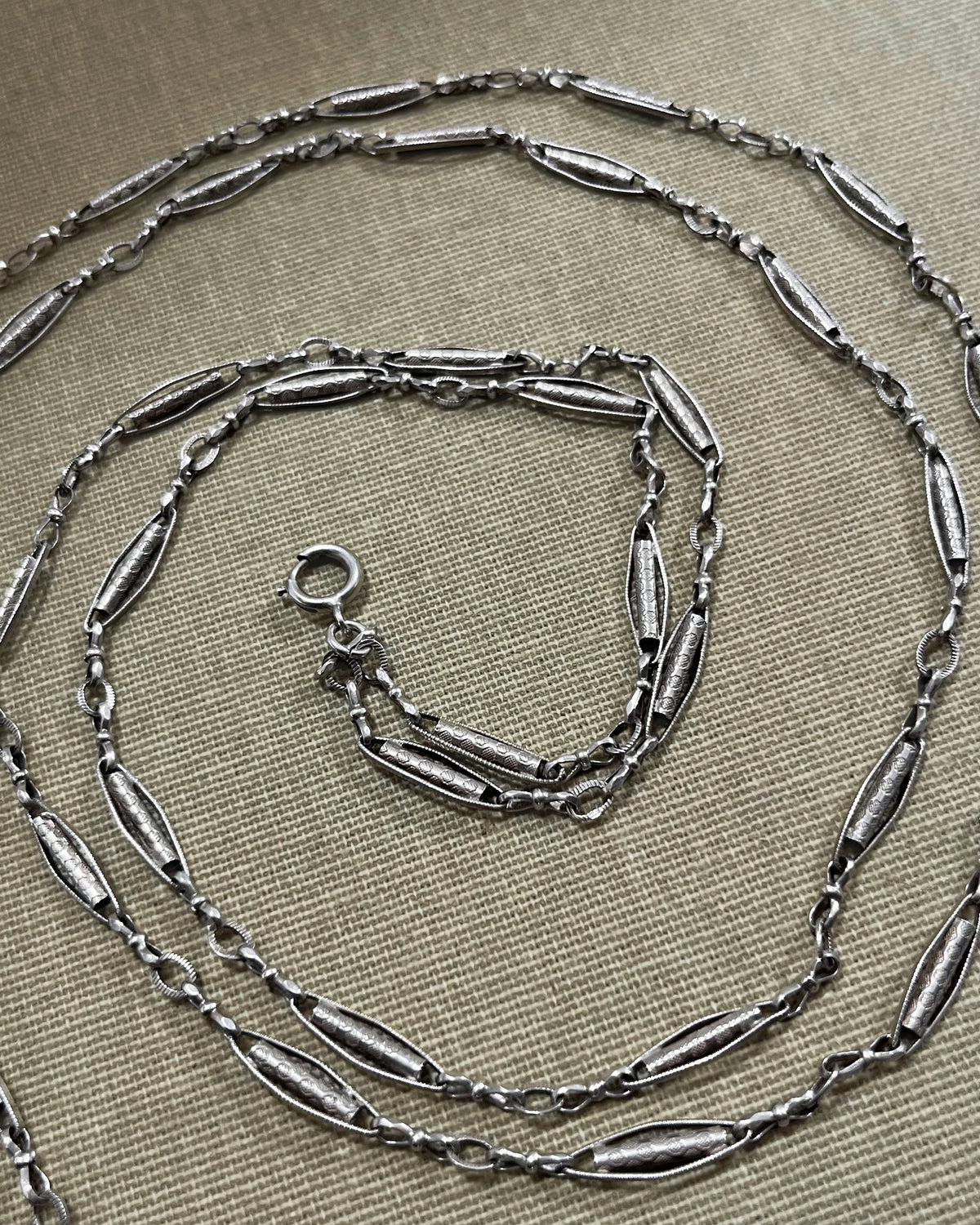 Belle Époque French antique silver sautoir necklace with decorative tubular, marquise and belcher links, arranged in a spiral on textured beige canvas.