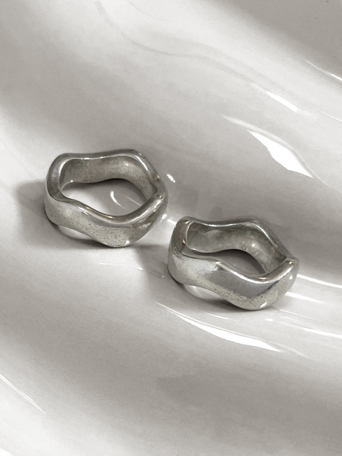 Two modernist vintage sterling silver wave rings separated on an undulating white tile, showing the individual curved forms and worn patina.