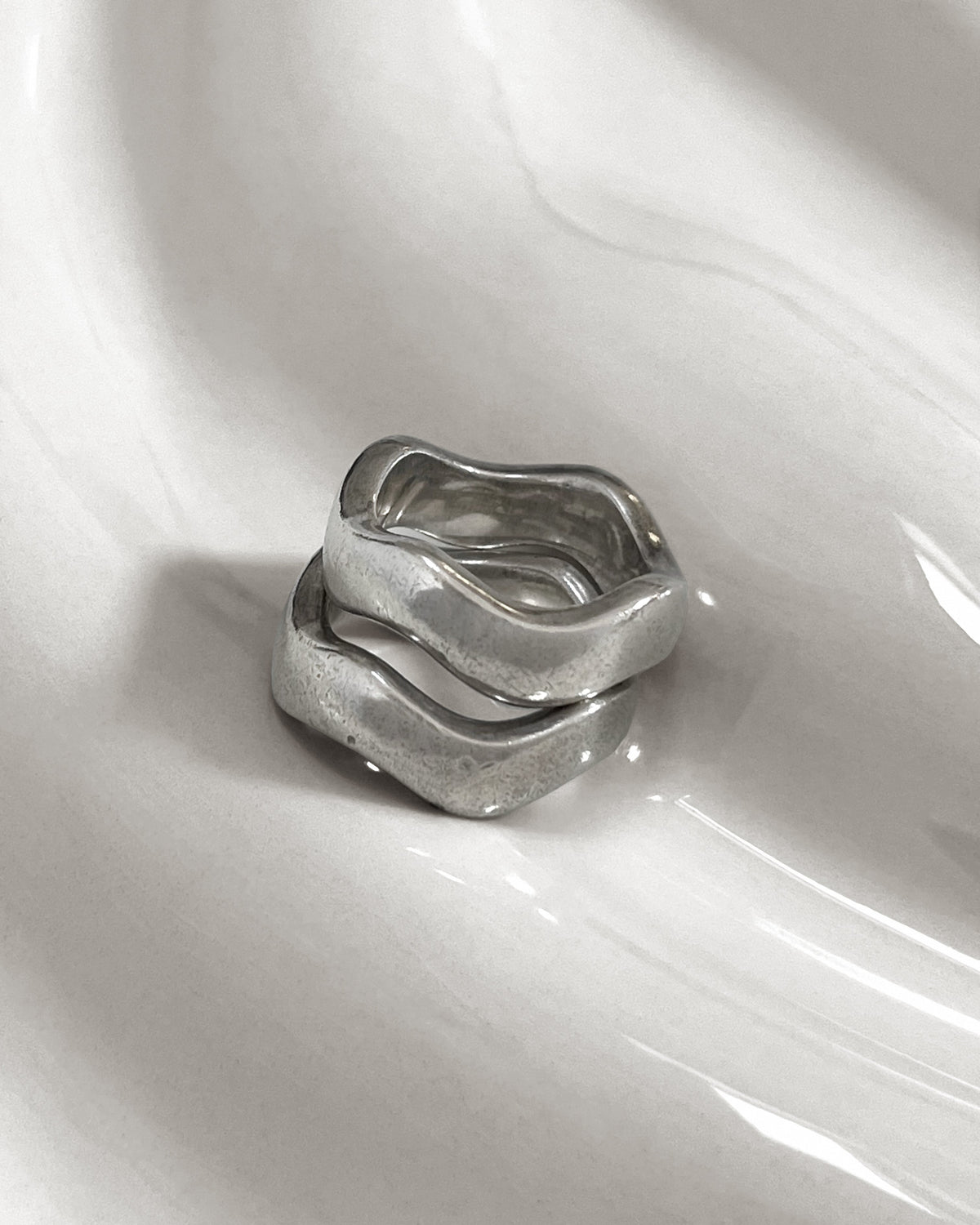 Two modernist sterling silver wave rings stacked together on an undulating white tile, showing their curved bands and softly worn patina.