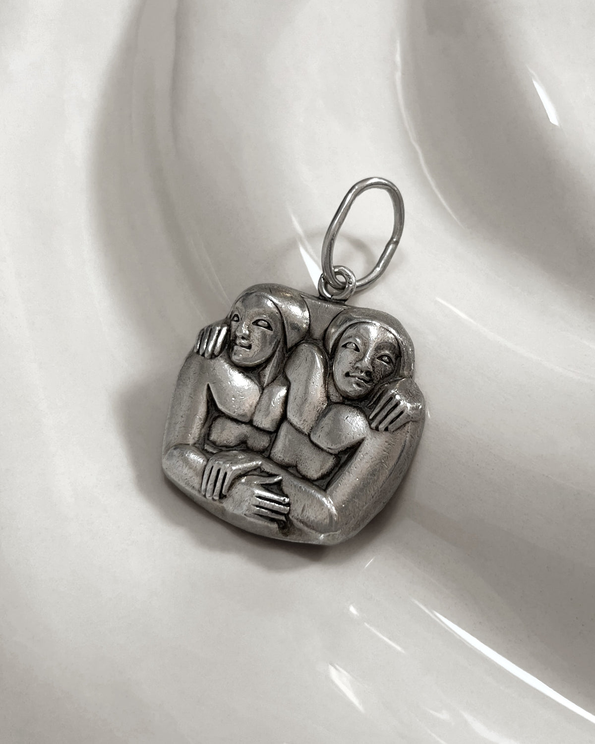 1940s French vintage sterling silver Gemini pendant with carved entwined twin figures, shown on a white undulating tile