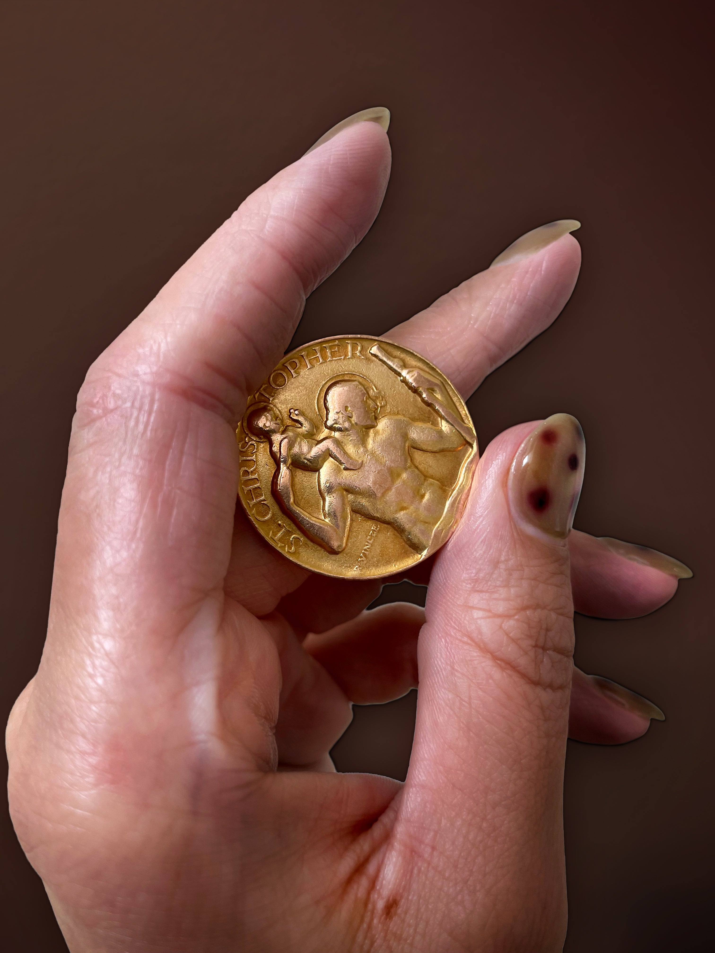 Hand holding a vintage 9kt yellow gold Saint Christopher medal by Paul Vincze against a brown background