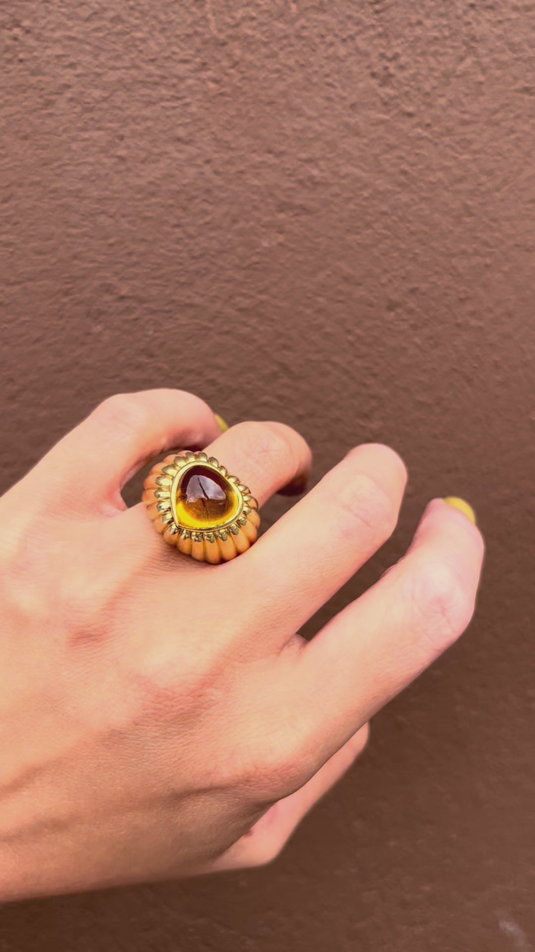 Video of a vintage chunky 18kt gold statement ring with a heart-shaped golden citrine cabochon and ribbed detailing, shown on a hand against a brown wall.